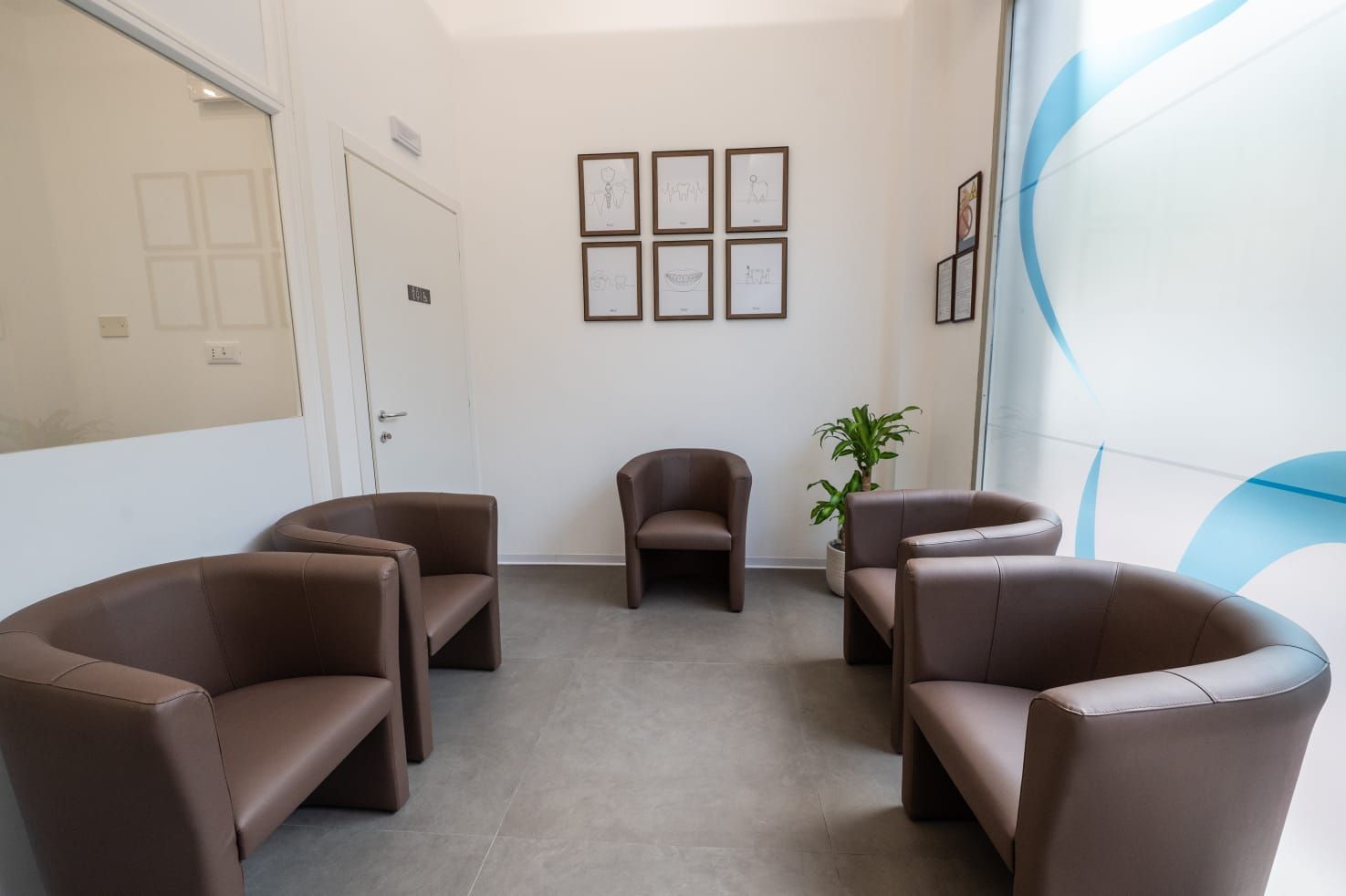 Una sala d'attesa con cinque poltrone marroni disposte a U, una piccola pianta verde e documenti incorniciati alle pareti.