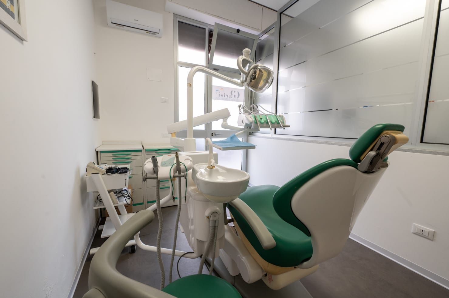 Una sala per trattamenti odontoiatrici sterile, dotata di poltrona odontoiatrica reclinabile verde e bianca, attrezzature mediche e lavandino.
