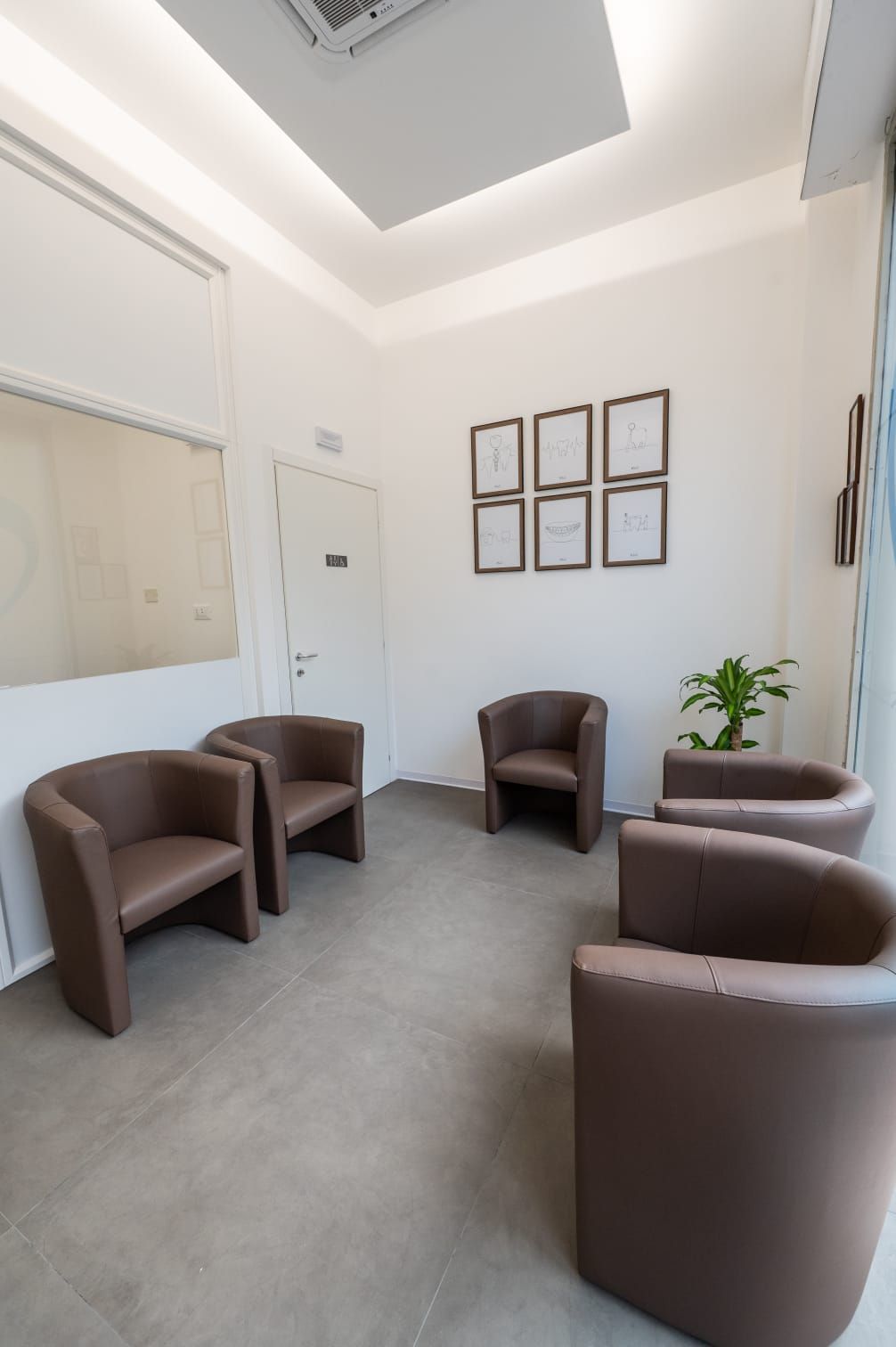 Una sala d'attesa con pareti chiare, pavimento in piastrelle grigie, cinque poltrone marroni disposte in cerchio e sei certificati incorniciati.