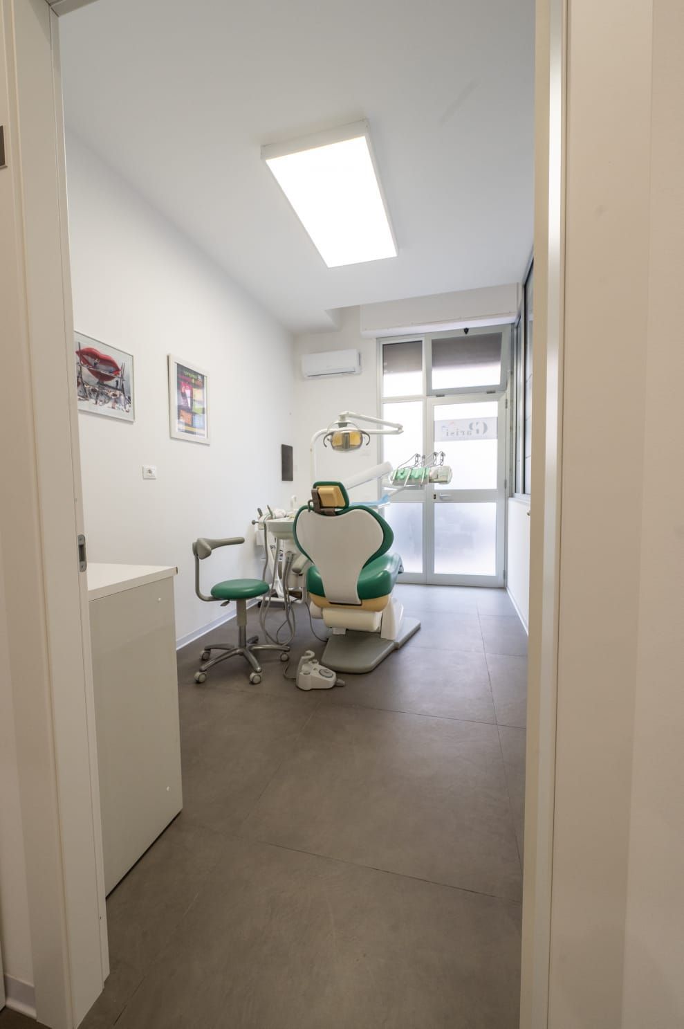 Una luminosa sala per visite odontoiatriche con una poltrona verde per il paziente, uno sgabello e un'ampia finestra in uno spazio pulito e bianco.