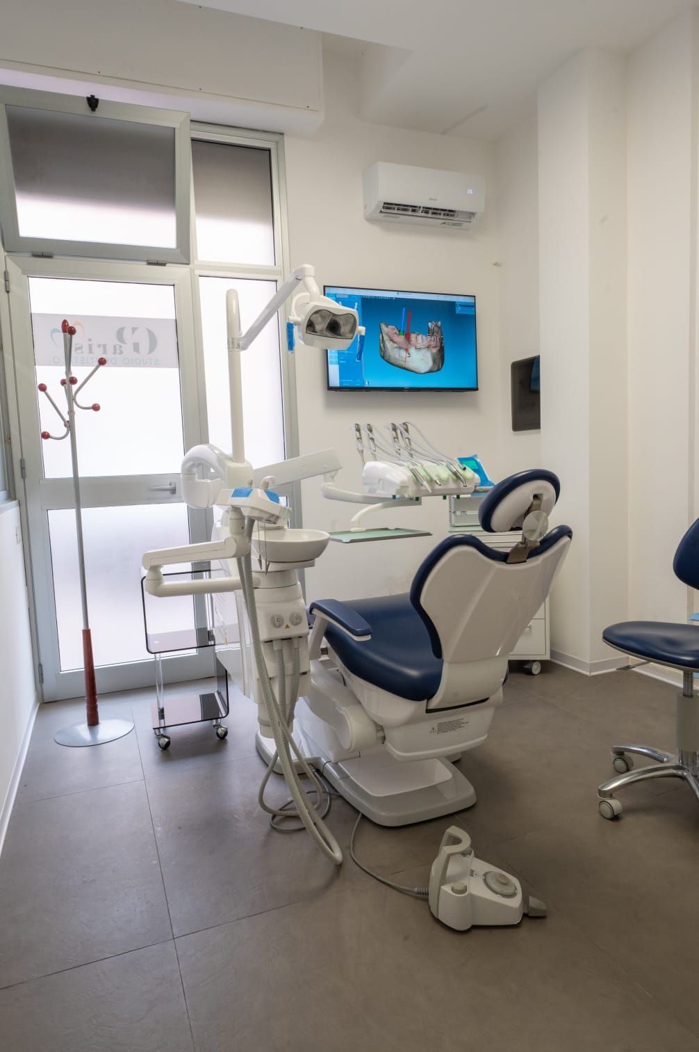 Una sala per visite odontoiatriche con una poltrona da trattamento blu e bianca, strumenti dentali e un monitor a parete che visualizza un'immagine.