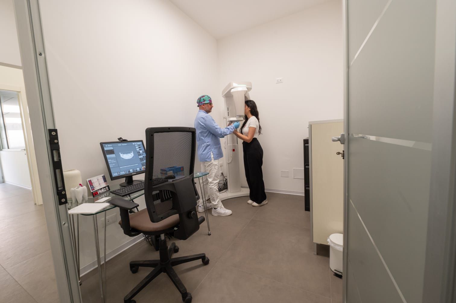 Un professionista sanitario assiste un paziente presso un apparecchio per la diagnostica per immagini dentale all'interno di uno studio medico dotato di postazione computer.