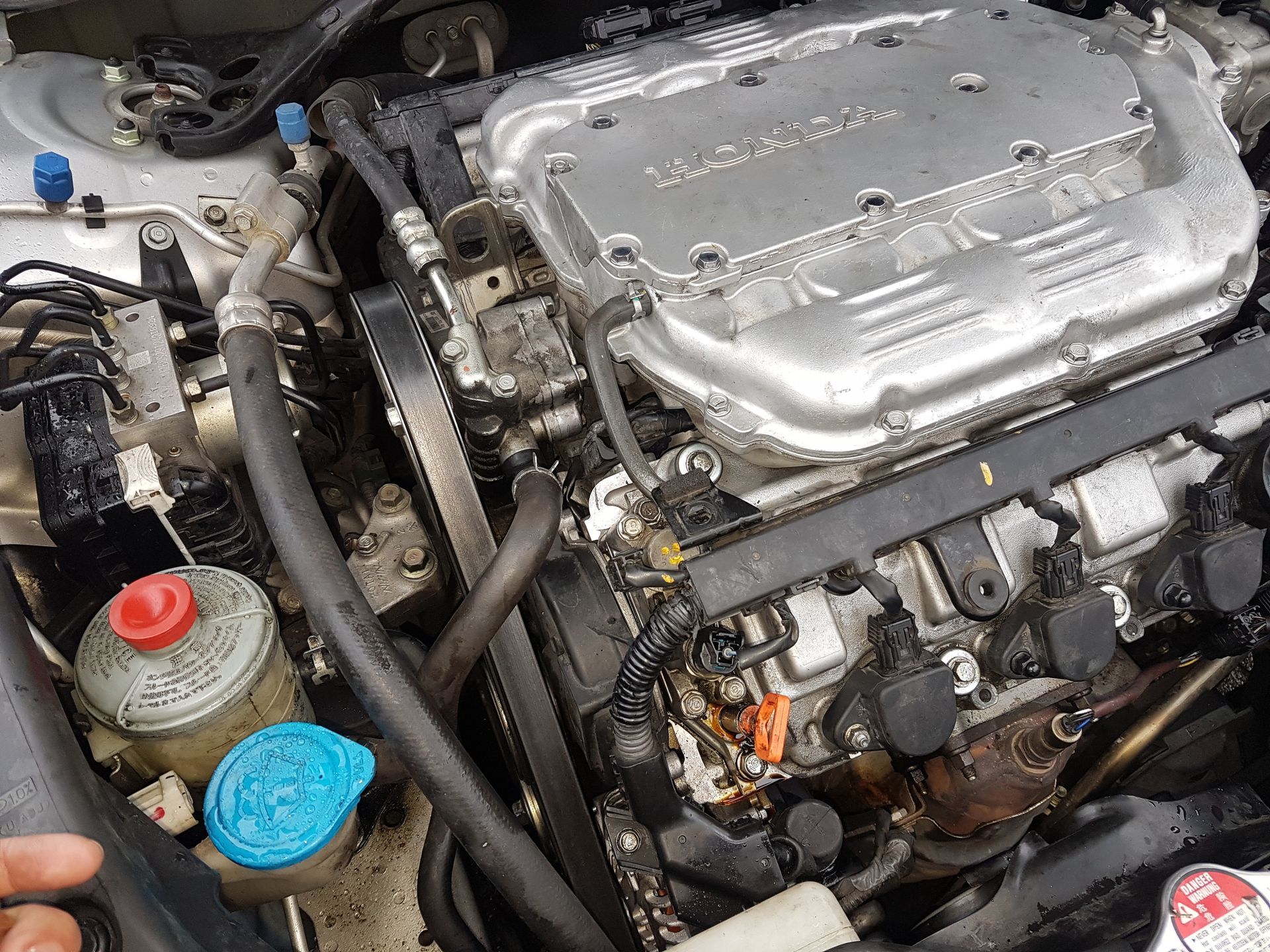 Close-up of a Honda V6 engine bay, showing the silver intake manifold, timing belt cover, and fluid reservoirs.