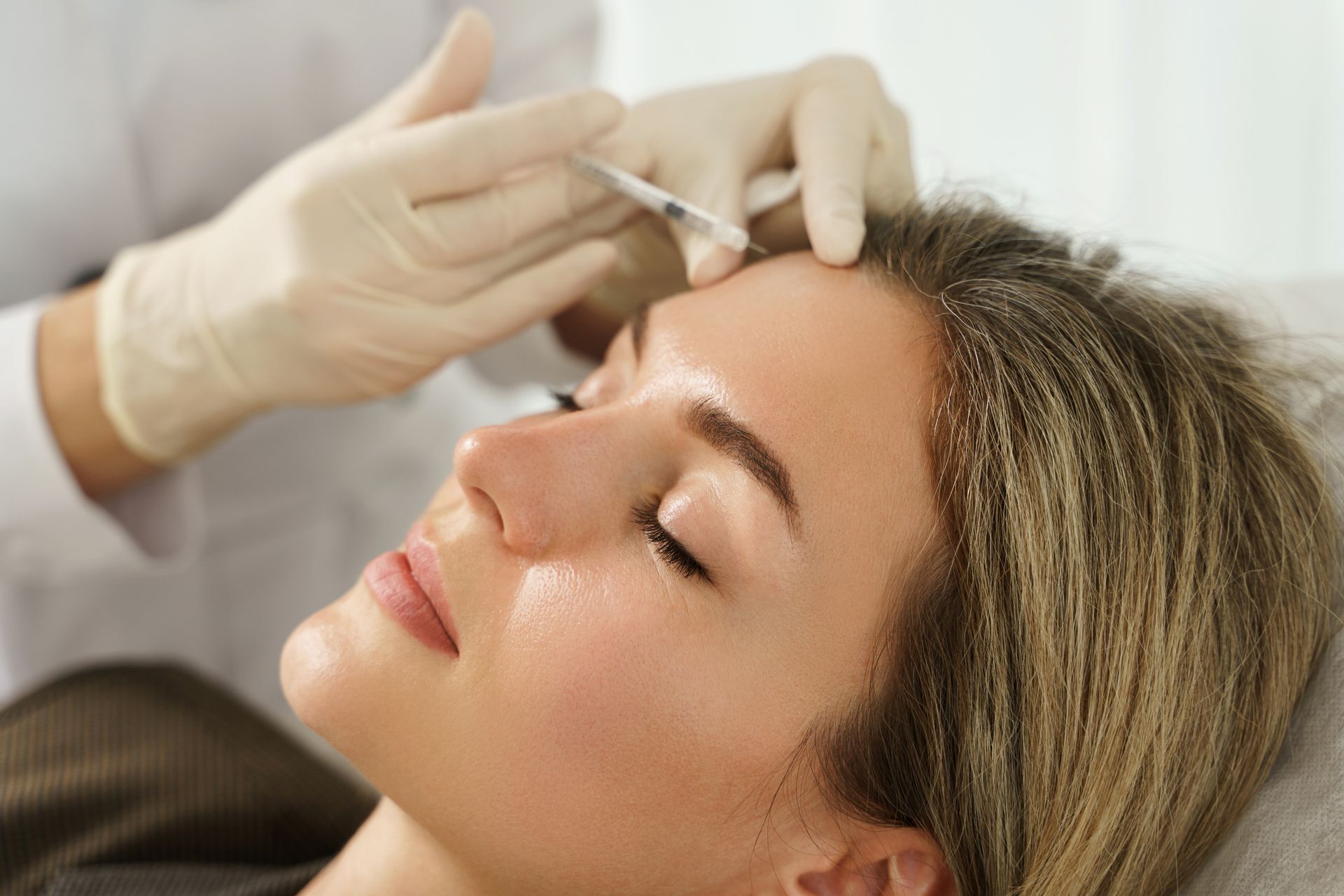 Gloved hands inject a woman's forehead while she lies relaxed on a treatment bed.