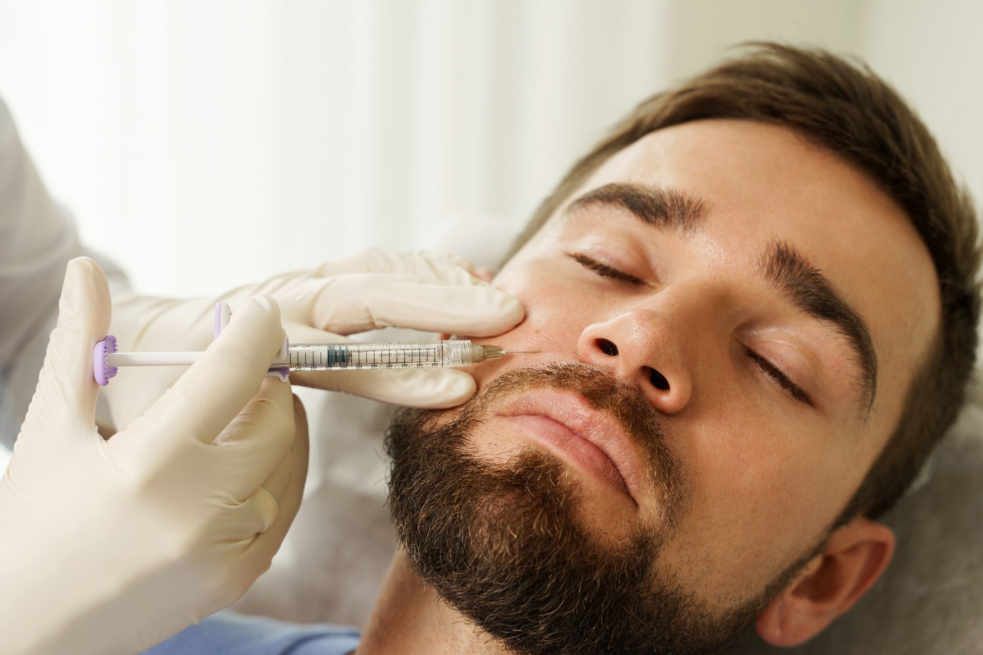 A practitioner in gloves performs a facial filler injection on a person's cheek.