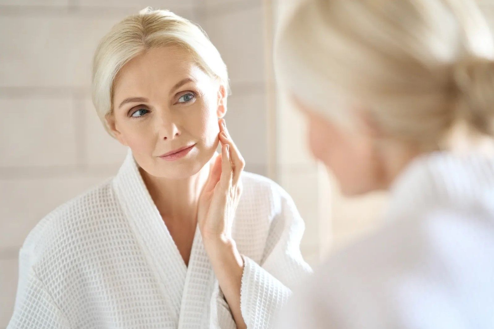 A woman in a bathrobe is looking at her face in the mirror.