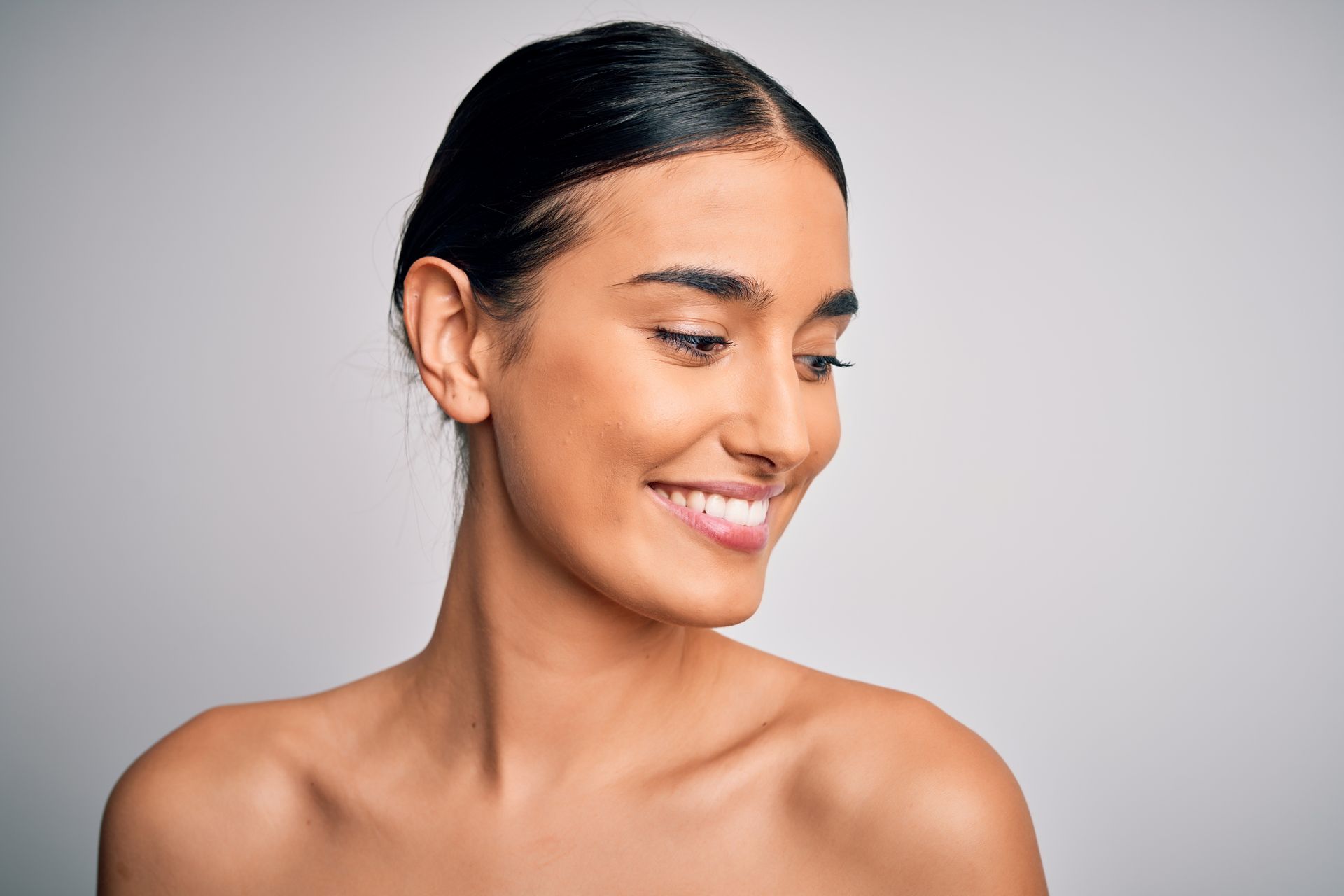A person with dark hair pulled back looks downward and smiles against a plain, light gray background.