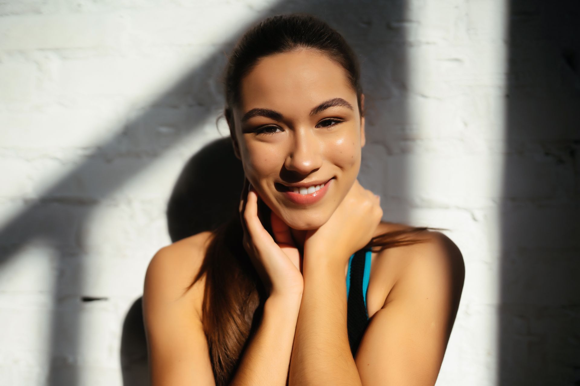 A woman is smiling with her hands on her face in front of a white brick wall.