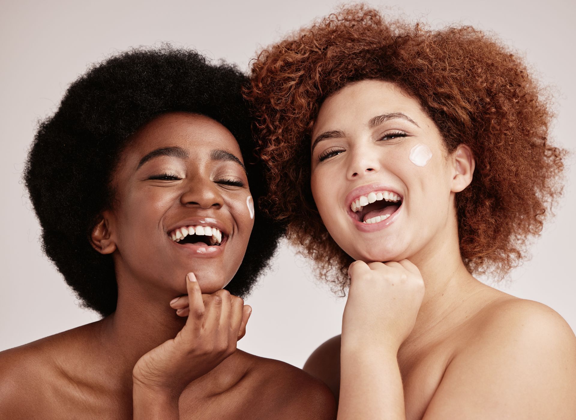Two women with applied skincare, smiling, holding chins.