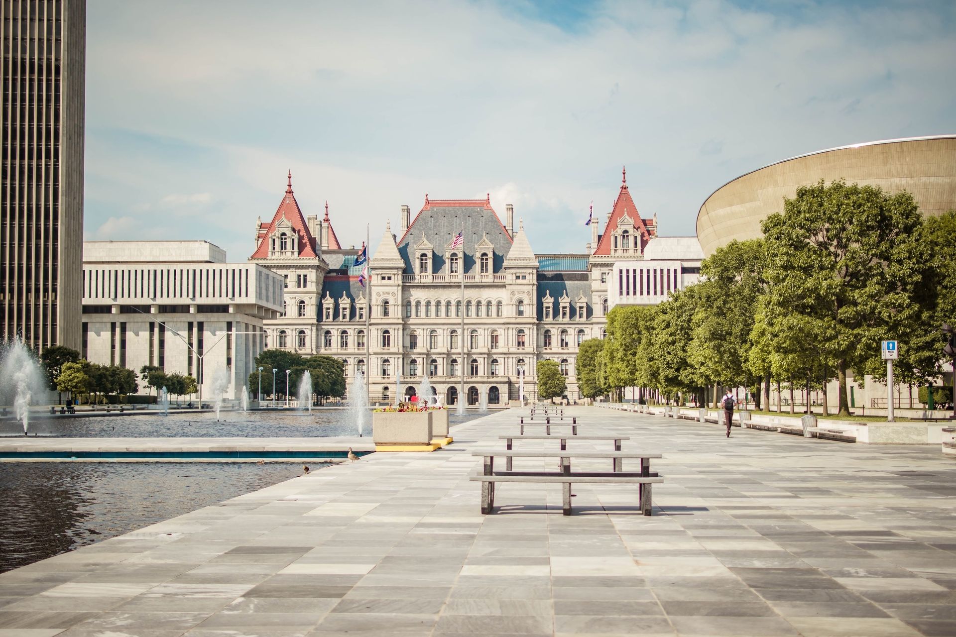 New York State Capitol building, Albany, with fountains, benches, and trees under a sunny sky.