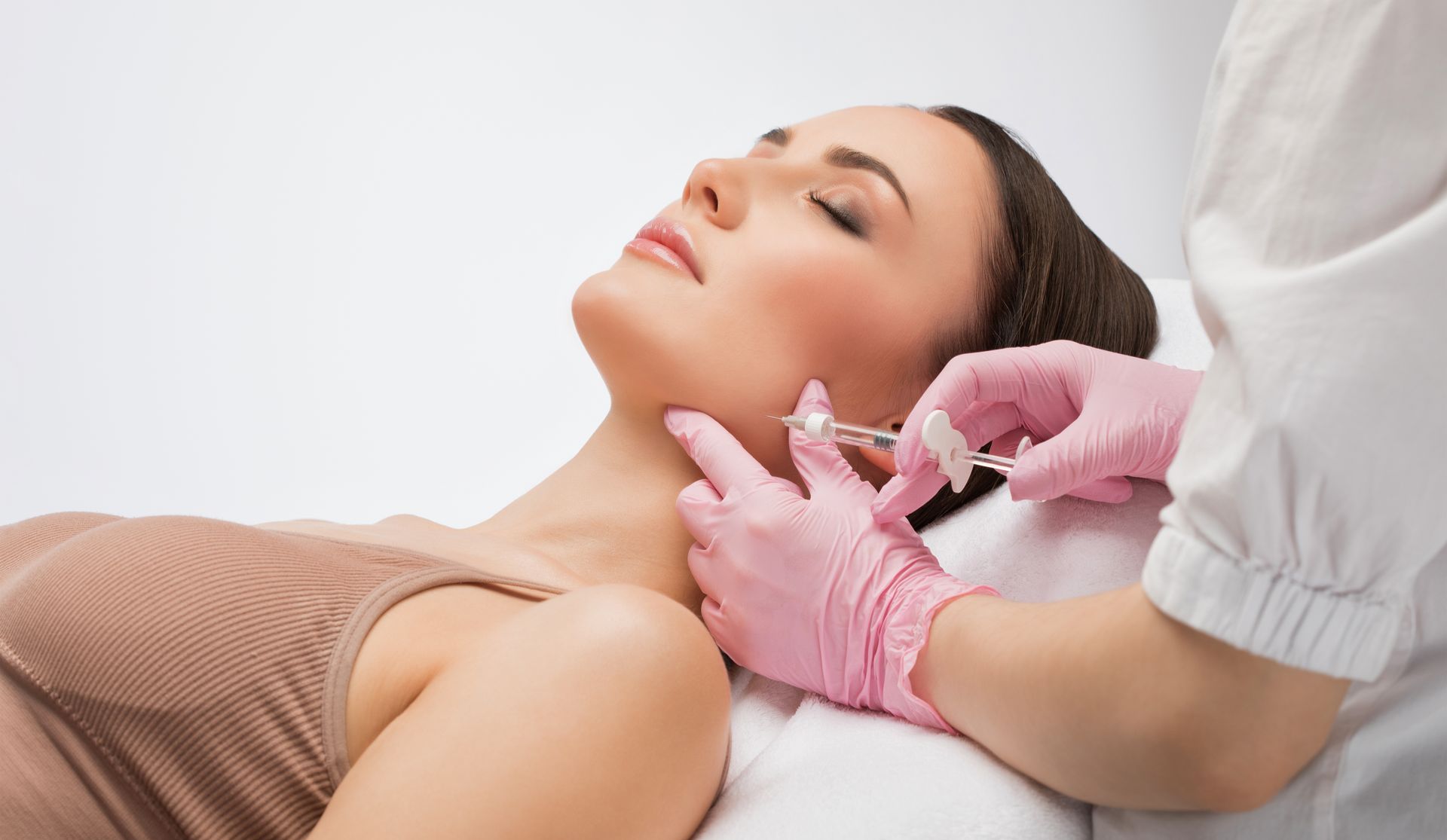 Aesthetic facial injection being administered by gloved hands to a reclining woman in a clinic