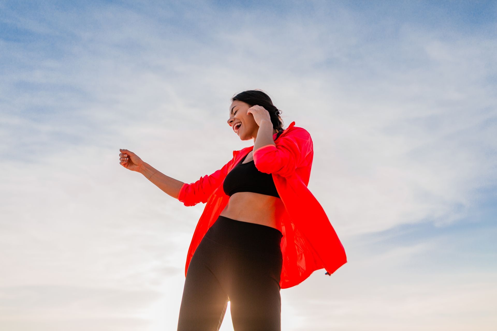 A woman in a red jacket and black pants is dancing on the beach.