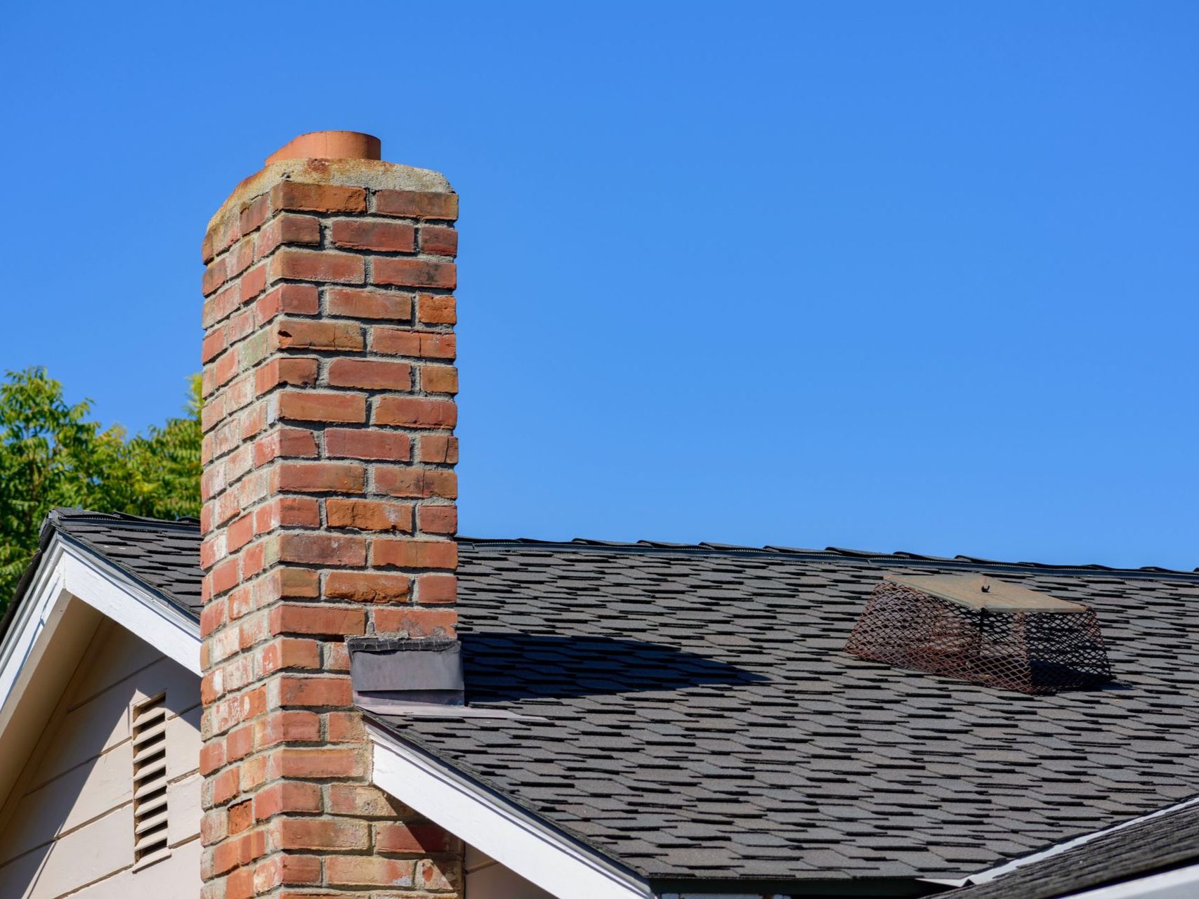 Brick chimney on a shingle roof against a clear blue sky.