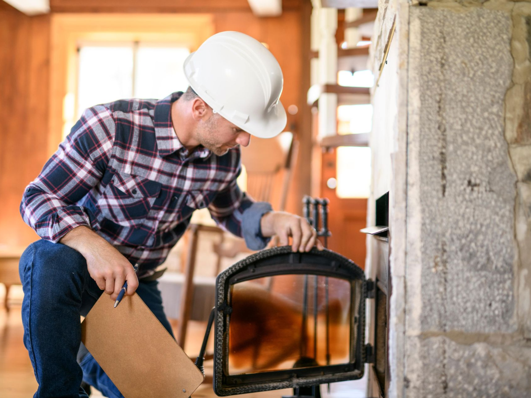 Man in hard hat inspecting fireplace. He holds a clipboard, wearing a plaid shirt and jeans.