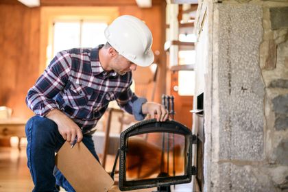 Man in hard hat examines fireplace, holding door, in a rustic, wood-paneled room.