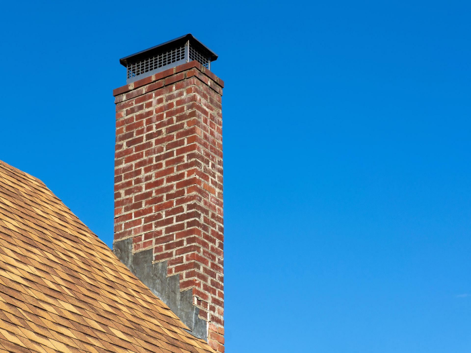 Brick chimney with a black cap on a roof against a blue sky.