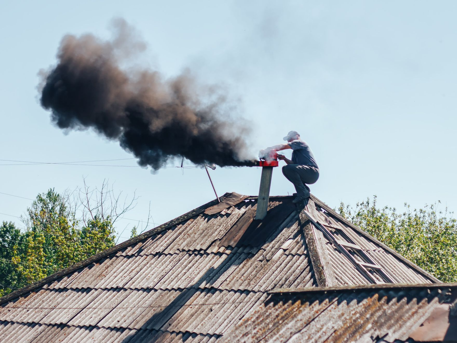 Person crouches on a roof next to a chimney emitting a large plume of black smoke against a blue sky.
