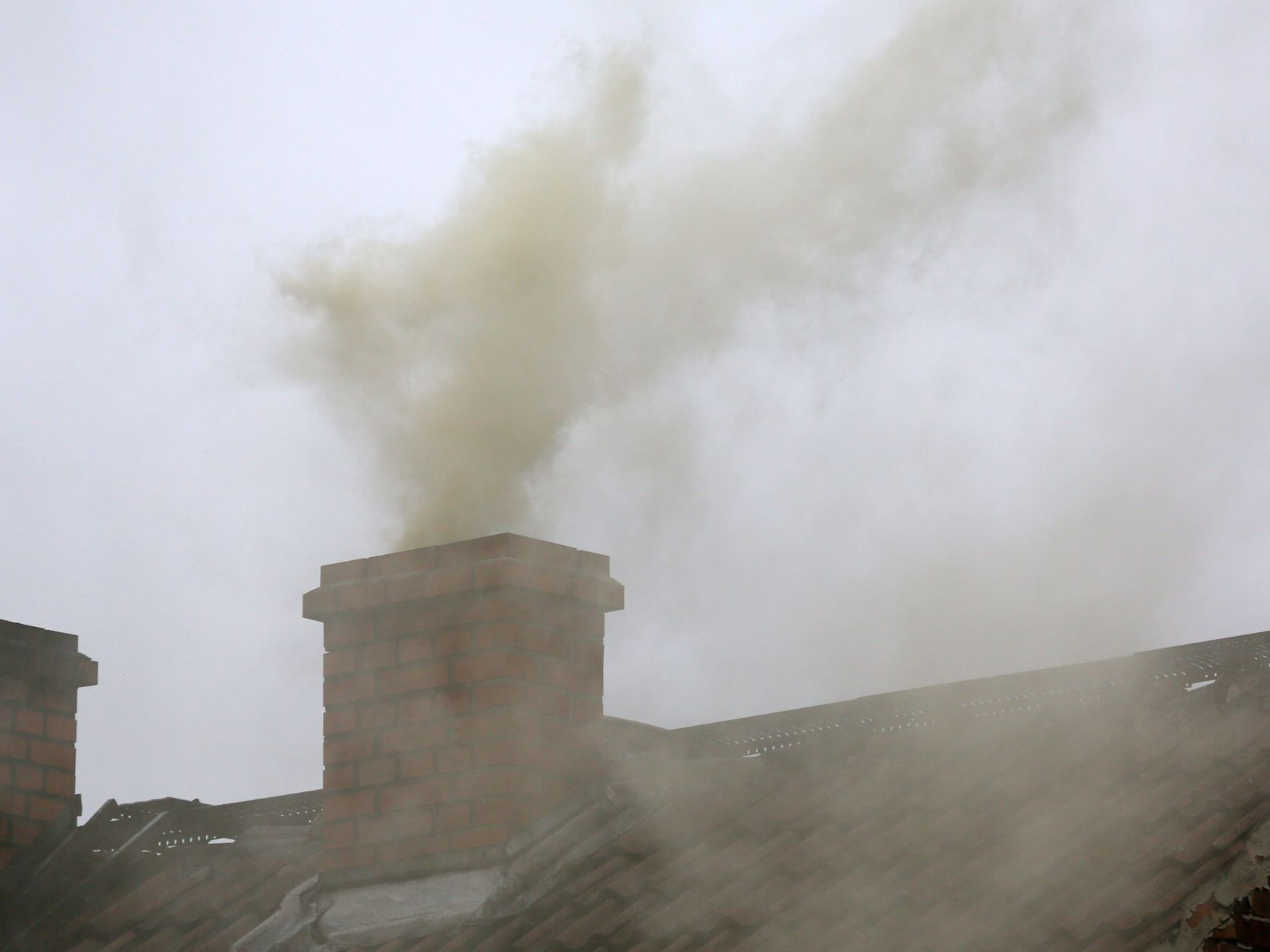 Smoke billowing from a brick chimney on a rooftop under a gray sky.