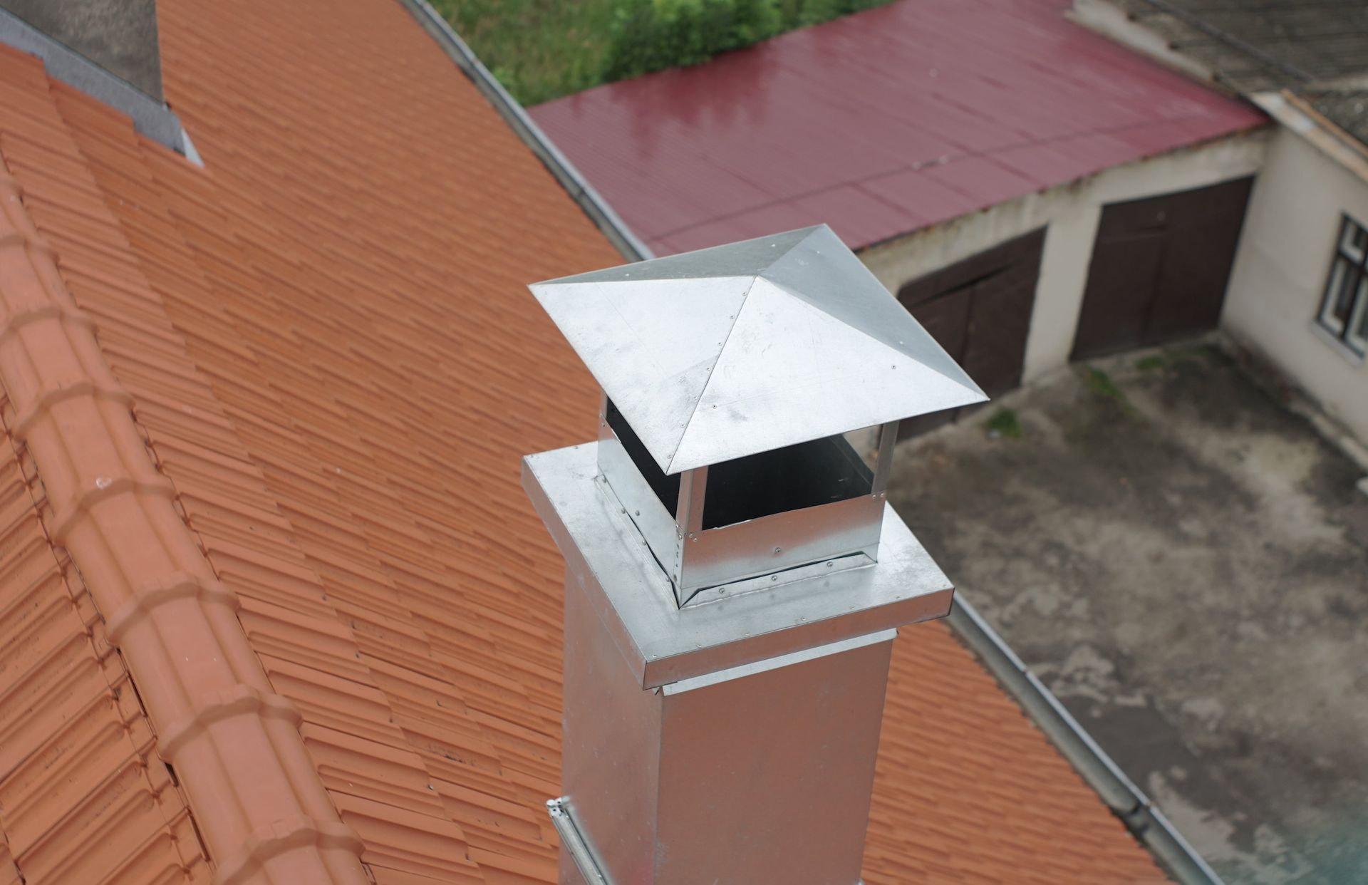 Metal chimney on a red tiled roof, topped with a square cap, outdoors.