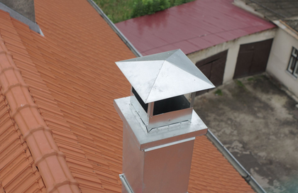 Metal chimney on a red tiled roof, topped with a square cap, outdoors.