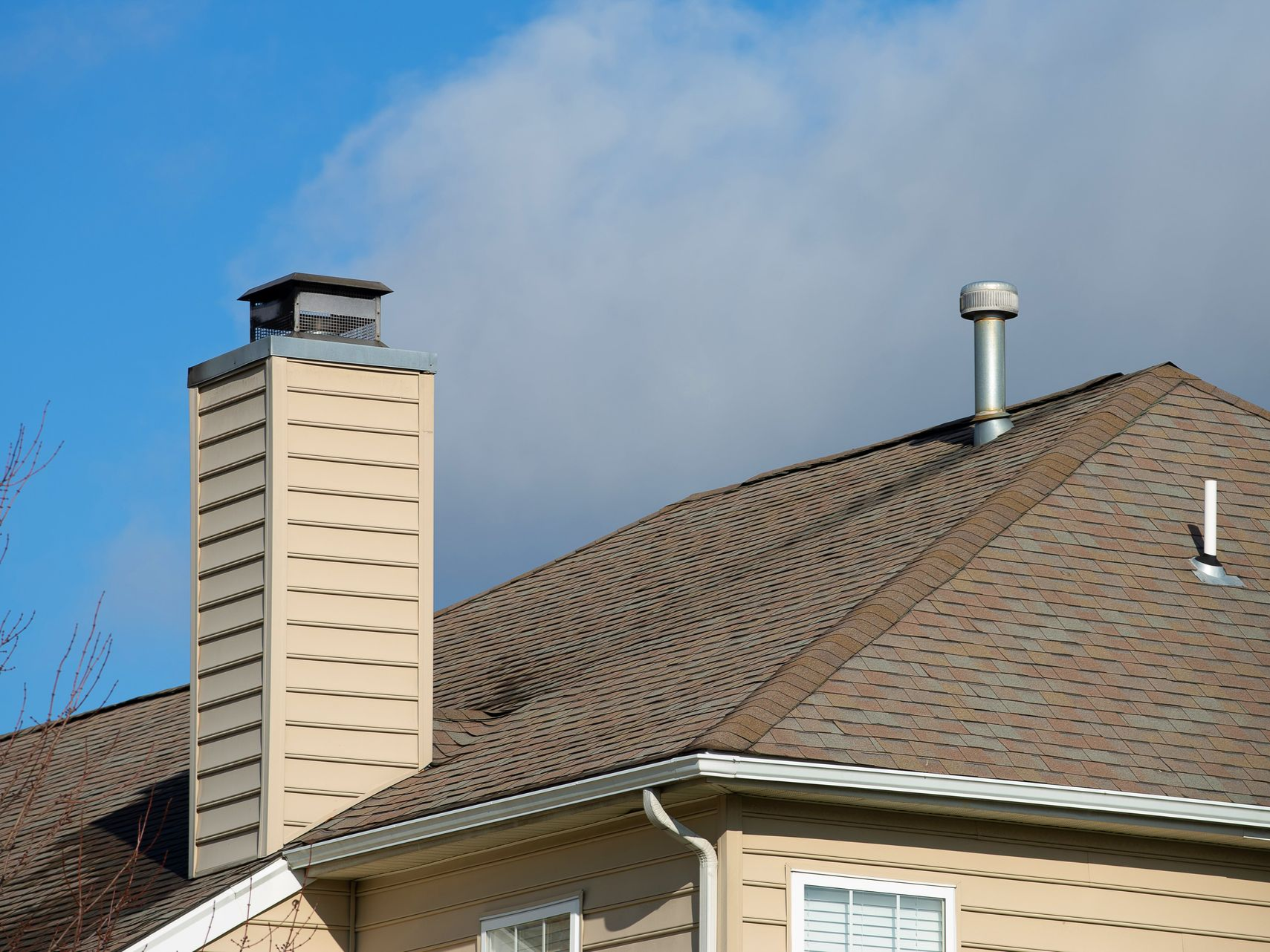 Tan chimney on a brown shingled roof against a blue sky with white clouds.