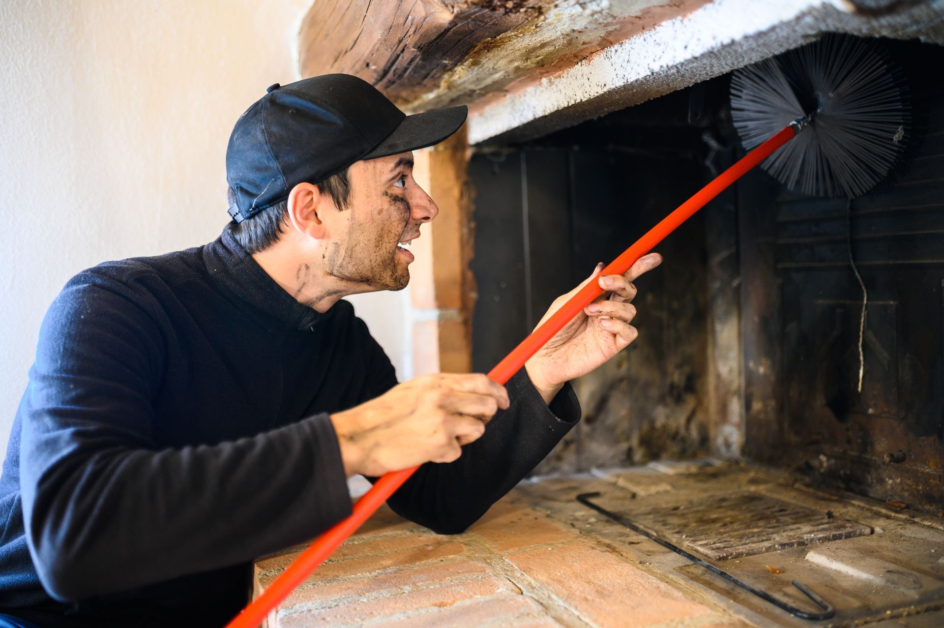 Man cleaning fireplace with brush. Black cap, orange handle, soot on face.