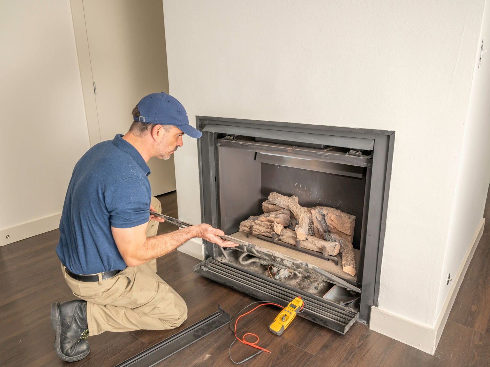 Man kneeling, examining fireplace. Tools on floor, neutral setting.