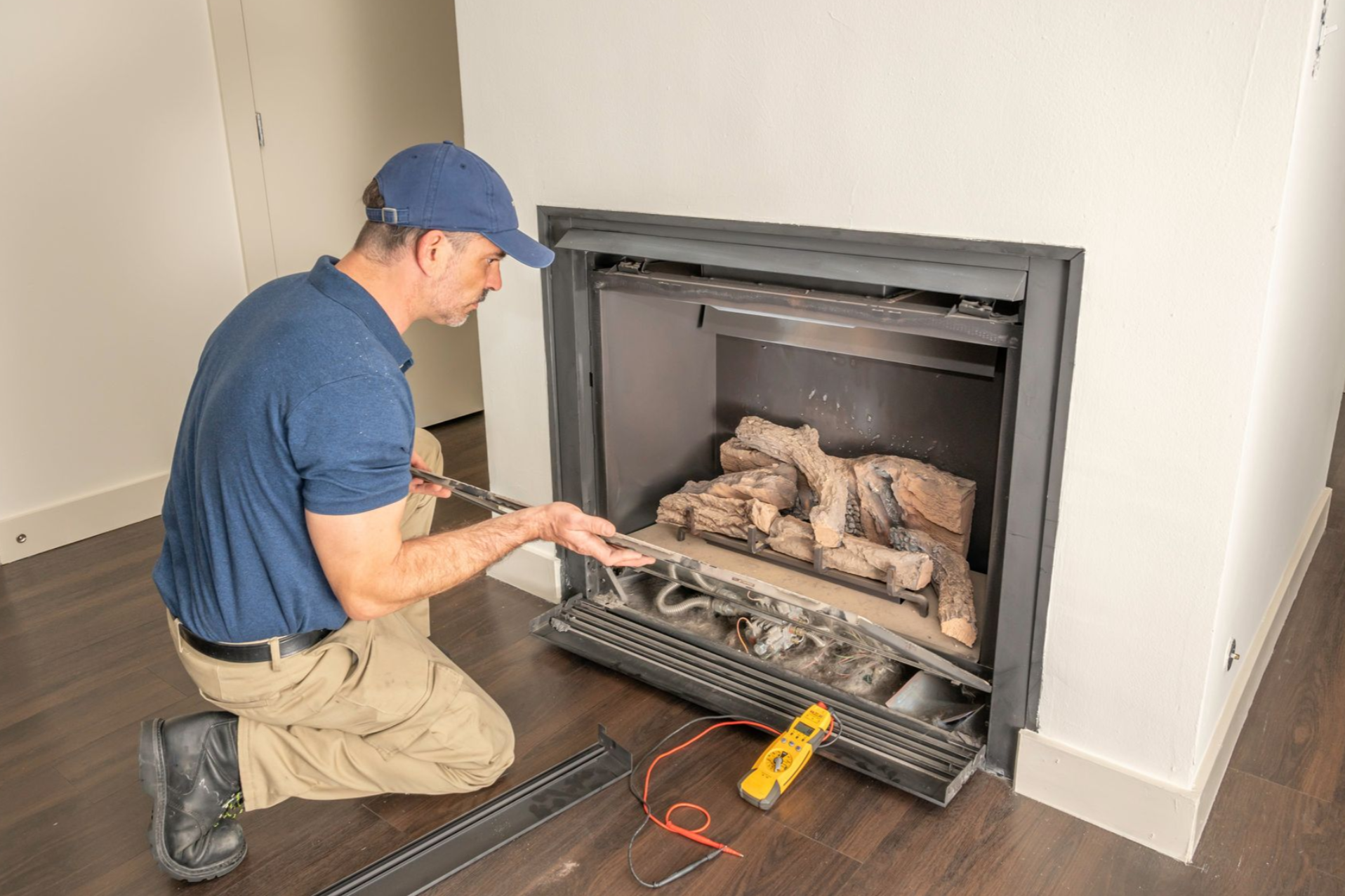 Man in blue shirt and cap servicing a fireplace, removing logs. Interior shot.