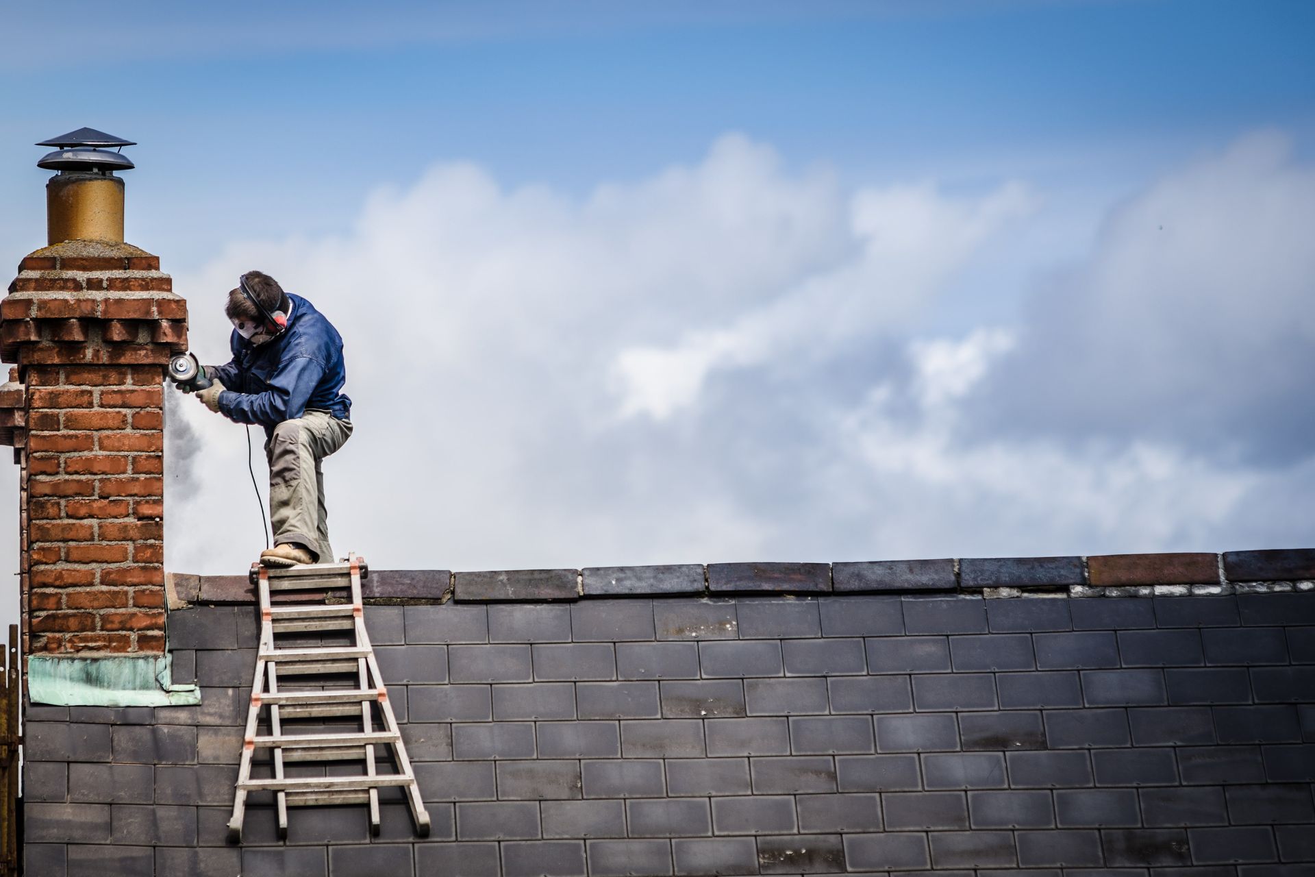 Person on ladder repairing a brick chimney on a dark-tiled rooftop under a cloudy sky.
