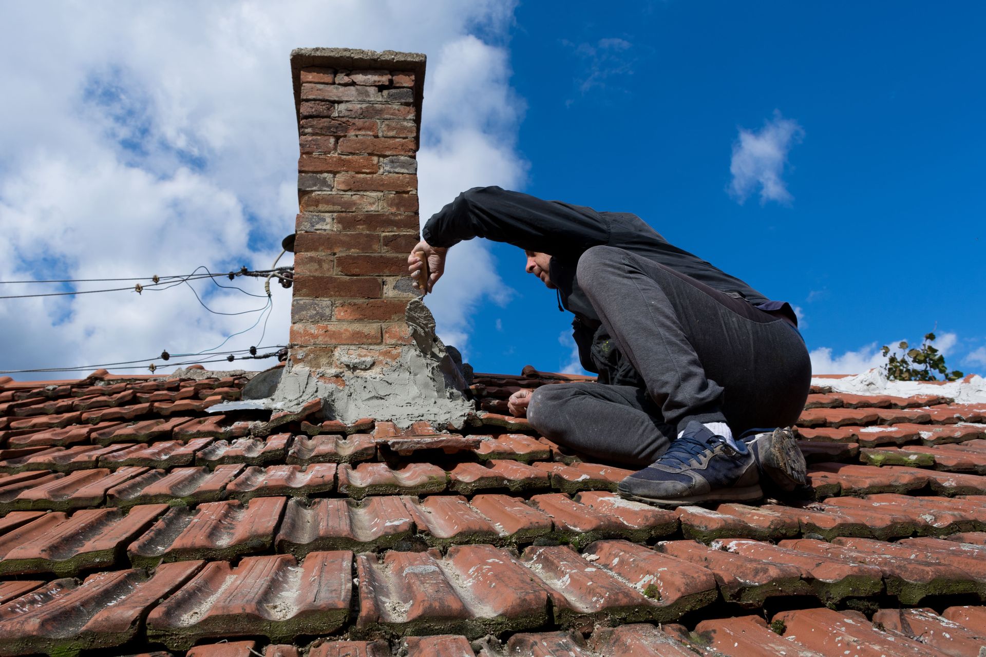 Person on roof inspecting chimney. Red tile roof, blue sky, brick chimney.