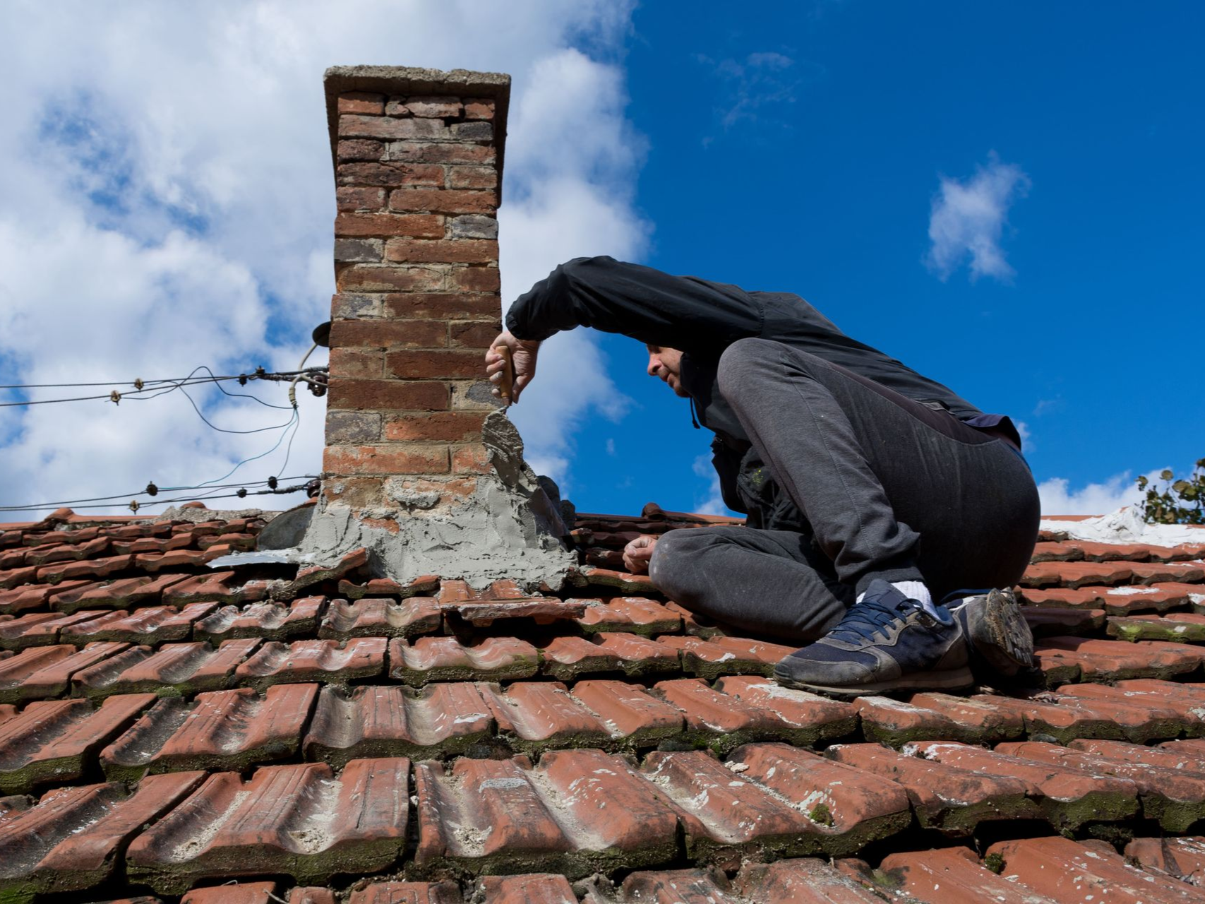 Person on roof near chimney, reaching toward it, blue sky.
