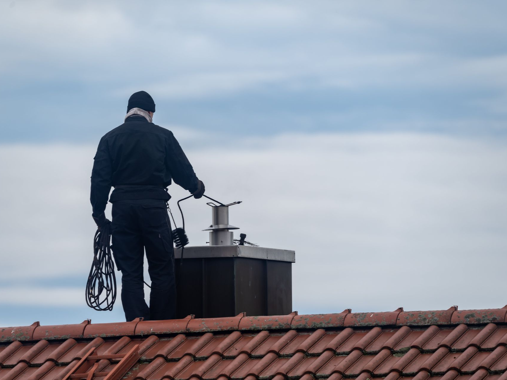 Chimney sweep on a rooftop, cleaning a chimney under a cloudy sky.