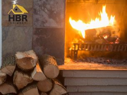 Fireplace with burning logs, firewood stacked nearby, HBR Chimney logo visible.