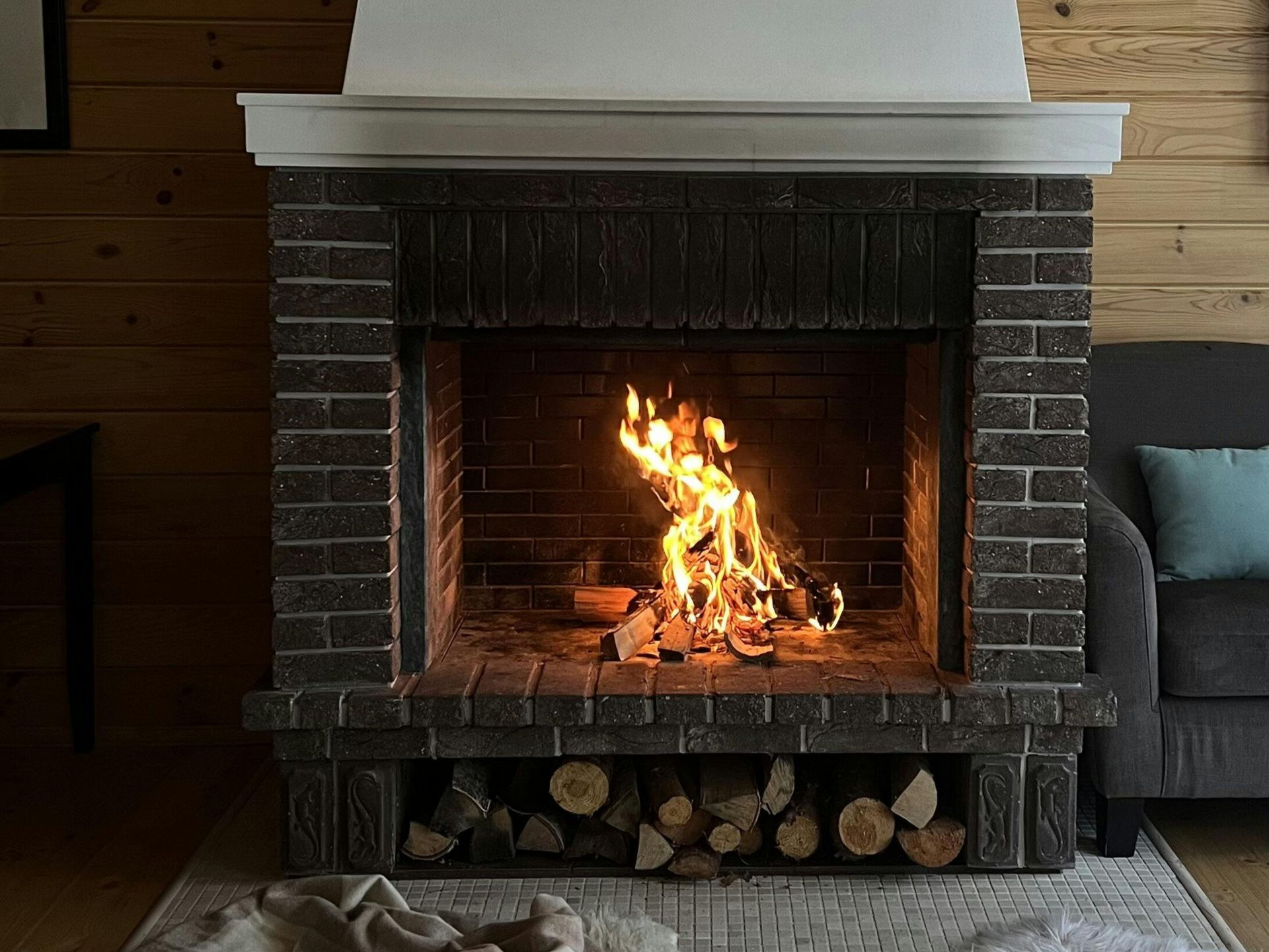 Fireplace with burning fire and stacked logs; brick facade, wooden wall.