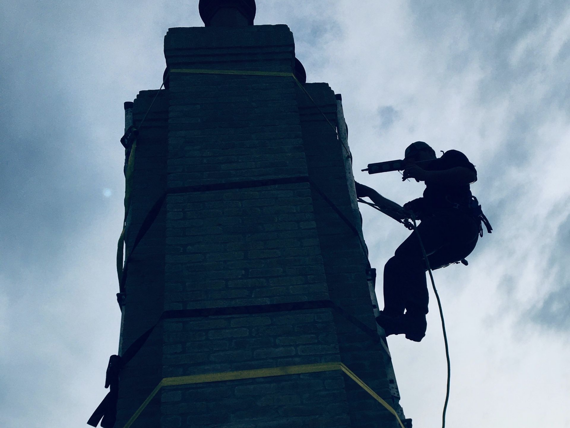 Silhouette of person rappelling down a tall, dark brick structure with cloudy sky in the background.
