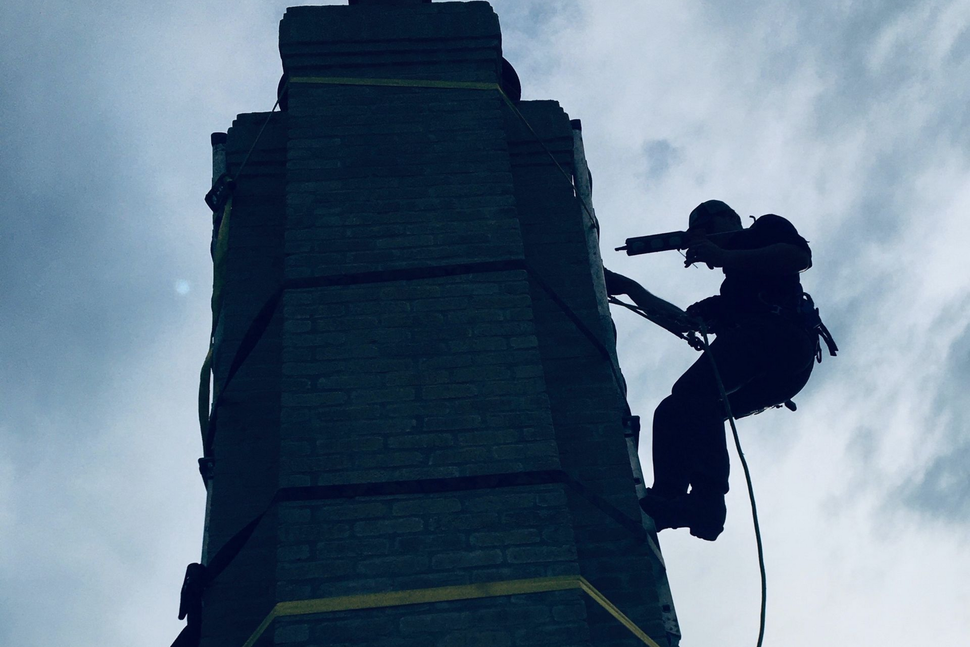 Person rappels down a tall, dark brick tower against a cloudy sky.