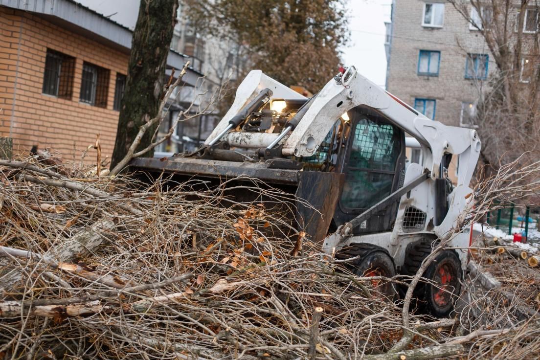 A white skid-steer loader clears a large pile of cut tree branches in front of a brick building.
