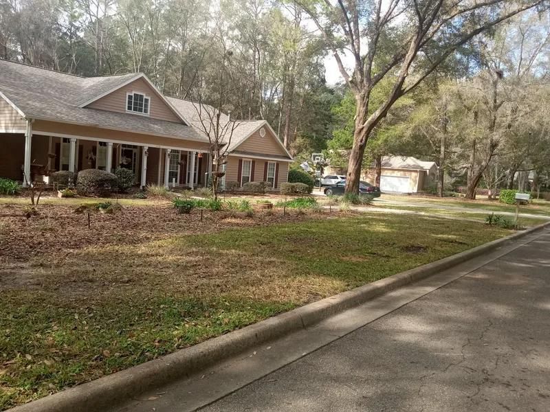 A single-story tan house with a front porch and columns sits in a grassy lawn.