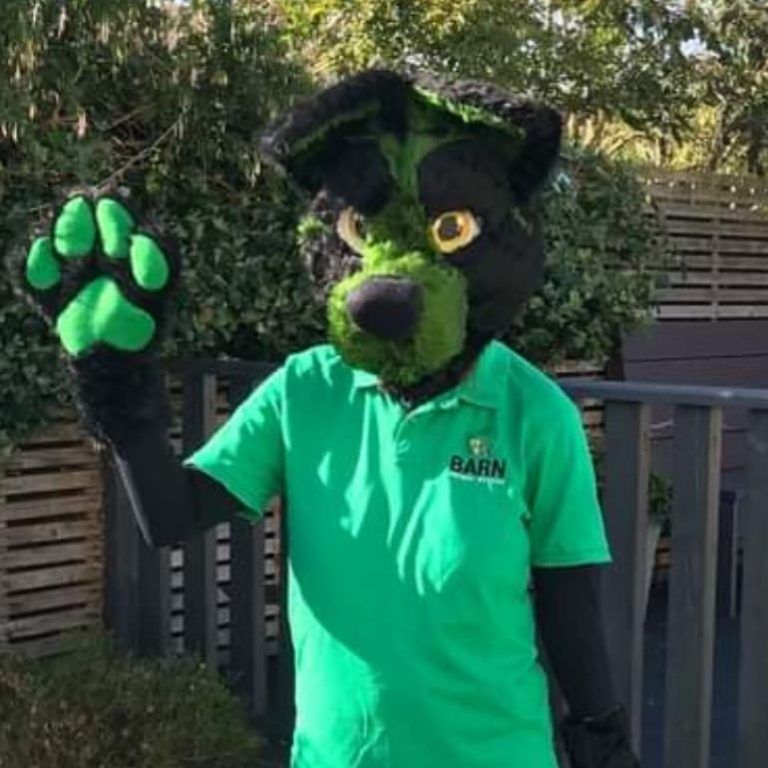 A black bear mascot wearing a green shirt and green paws is waving.