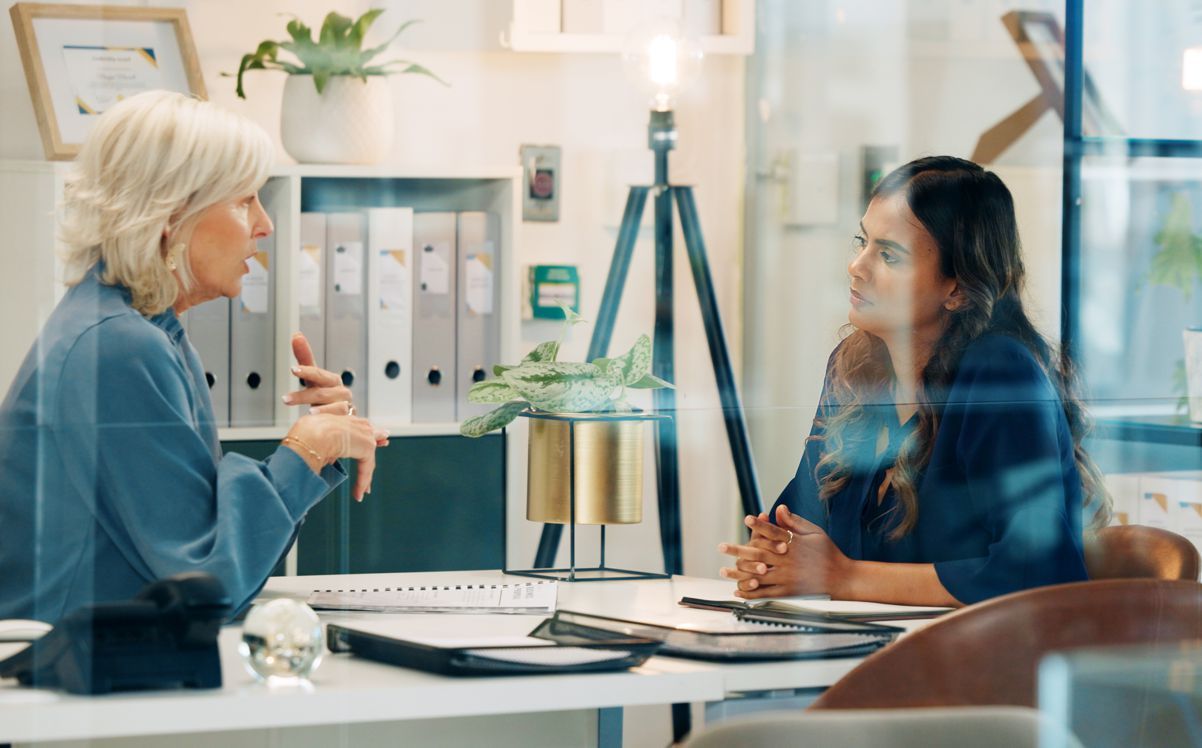 Two women are sitting at a desk having a conversation.