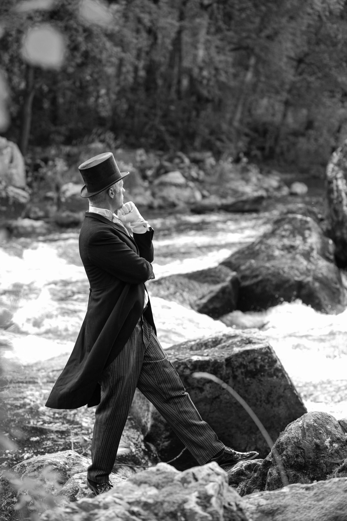 Silinterihattu ja muodollinen frakki päässä oleva henkilö seisoo kallioilla kuohuvan joen varrella ja lepää käsi leuallaan.