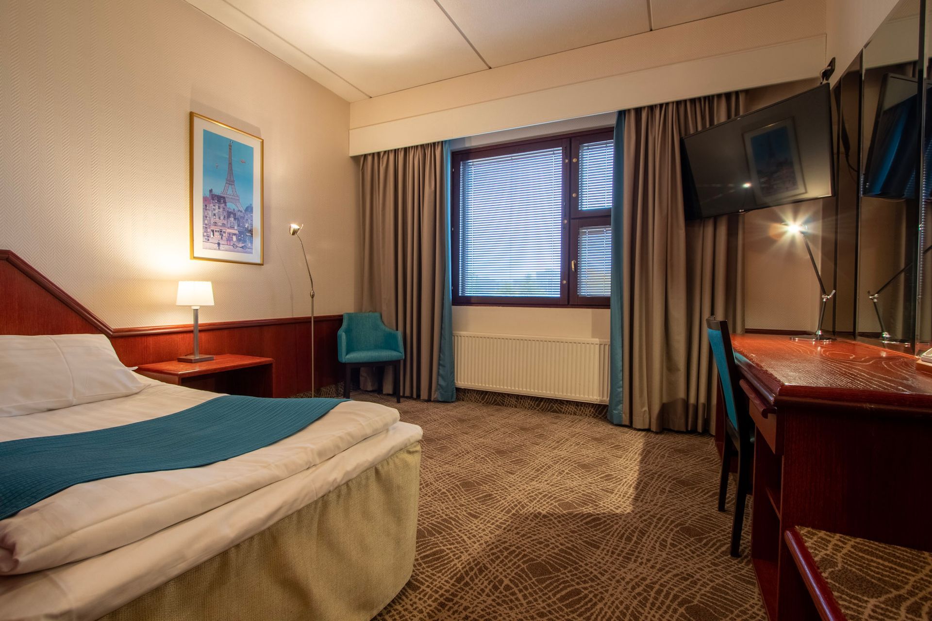 Hotellihuone, jossa on siististi pedattu sänky, puinen työpöytä ja siihen kiinnitetty televisio, sinivihreä tuoli ja ikkuna verhoineen.