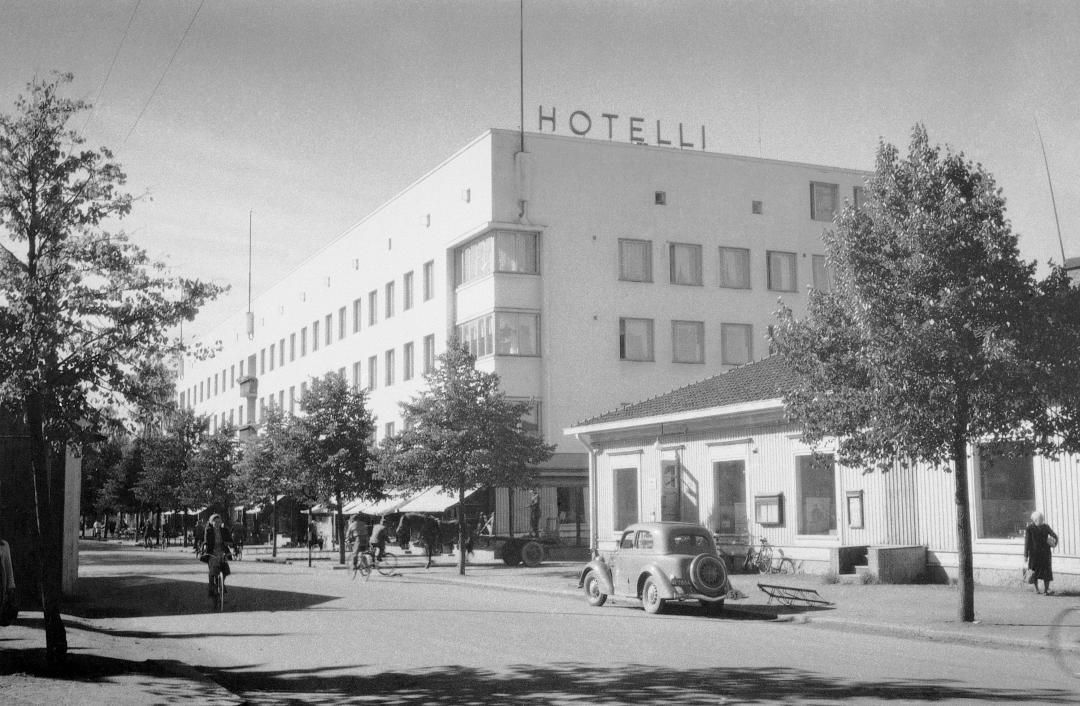 A historic black-and-white photo of a multi-story hotel building with "HOTELLI" sign, trees, and a car on a street.