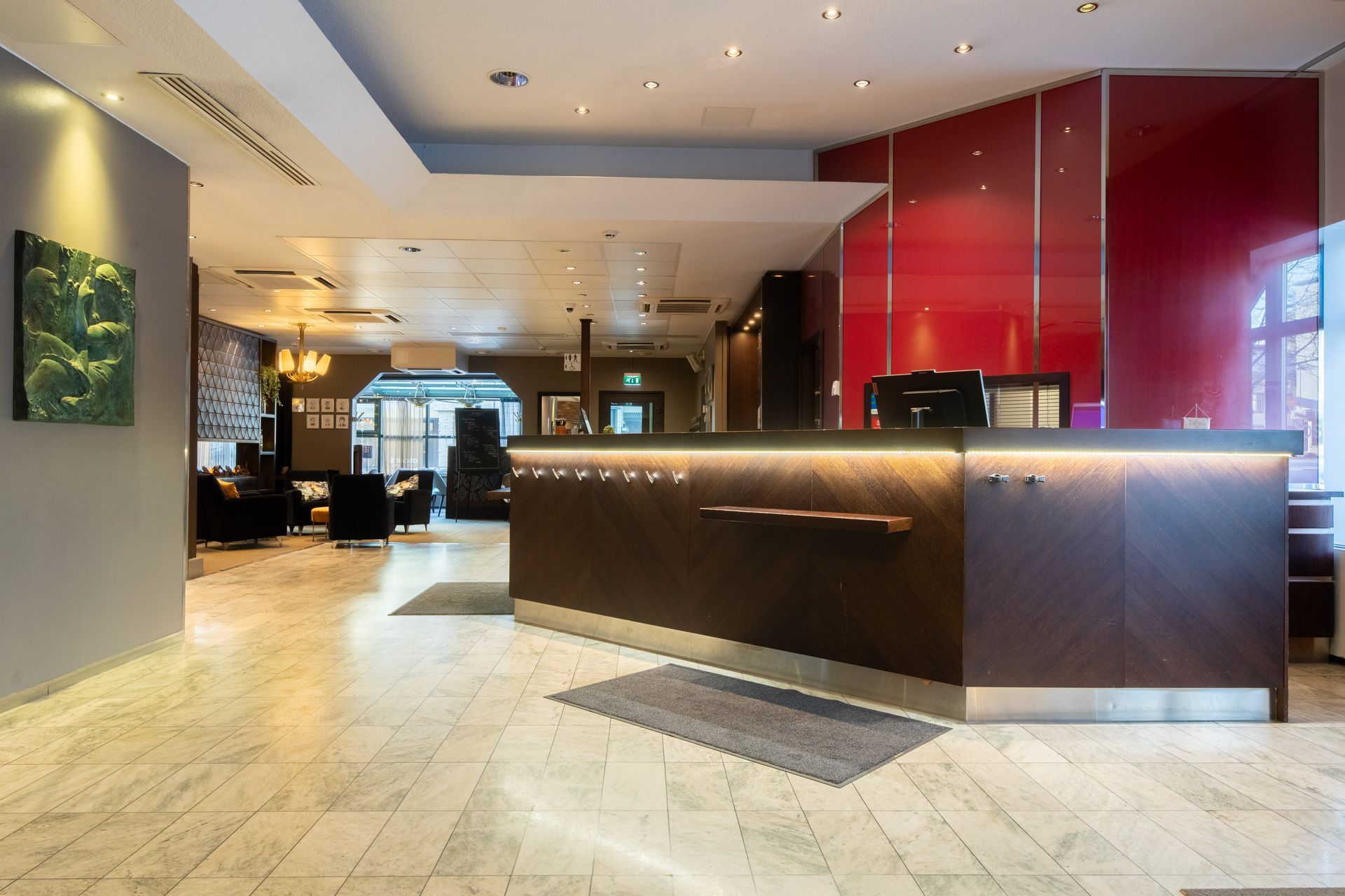A hotel reception area with a dark wood desk, marble flooring, and a bright red accent wall behind the front counter.