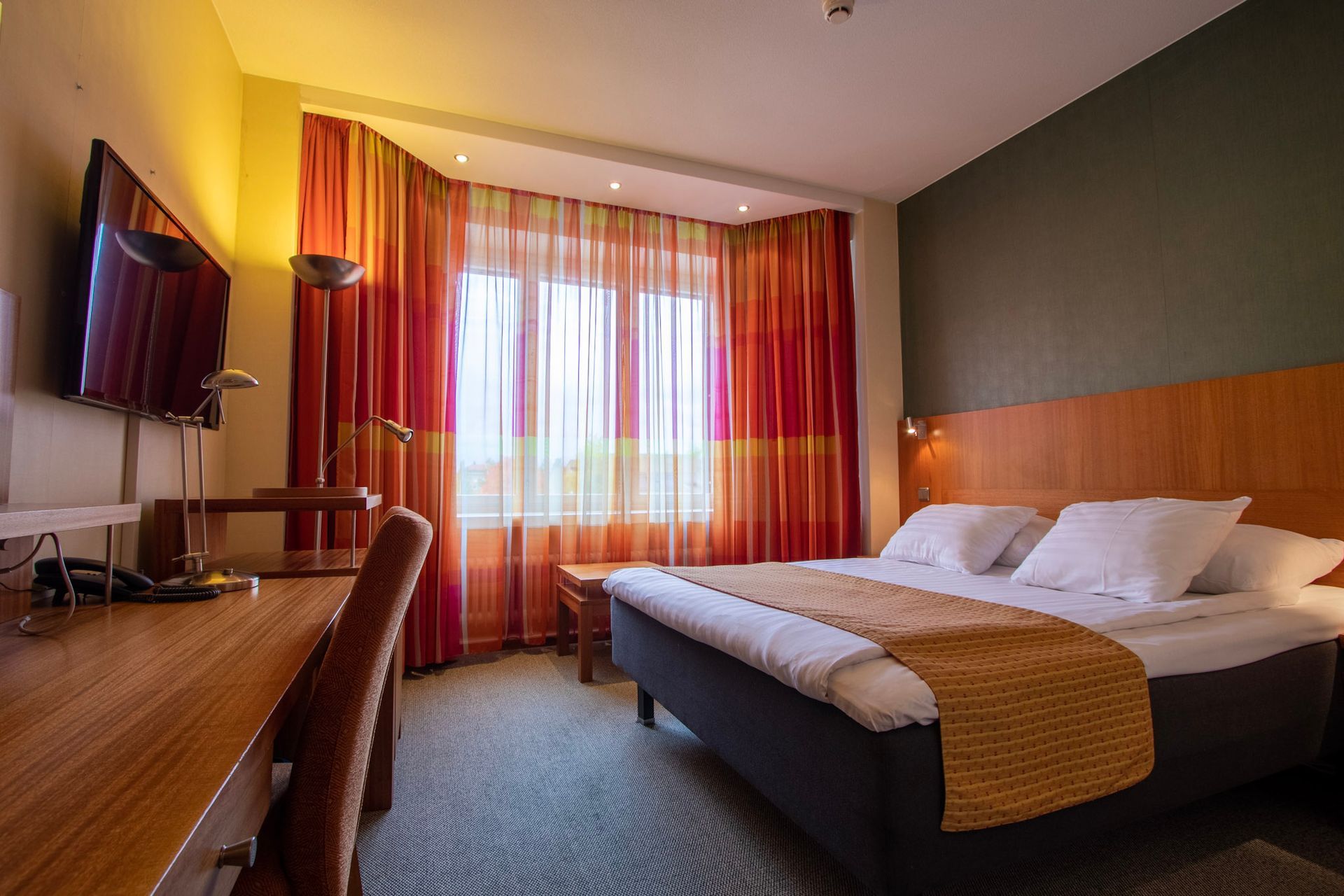 Hotellihuone, jossa on sänky valkoisine lakanoineen, työpöytä ja tuoli sekä suuret ikkunat kirkkaanpunaisine ja oransseine verhoineen.