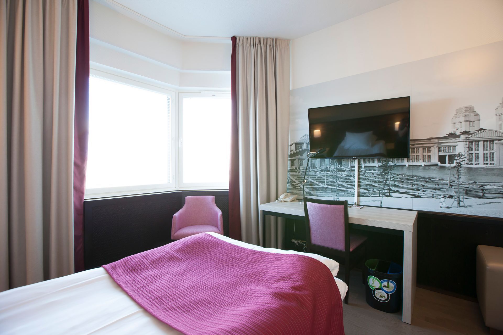 Kirkas hotellihuone, jossa on sänky magentanvärisellä peitolla, työpöytä tuolilla ja seinälle kiinnitetty taulutelevisio.