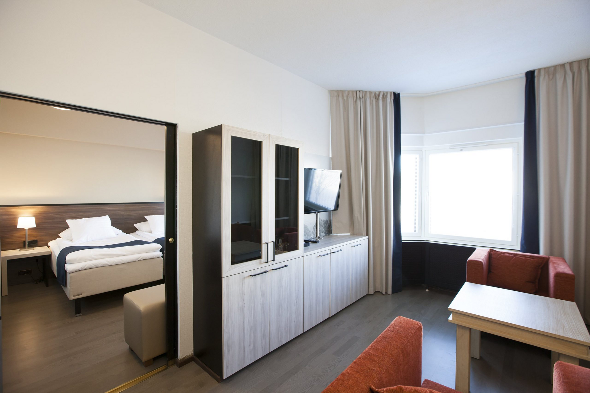 Hotellisviitti, jonka viereiseen makuuhuoneeseen näkyy oviaukko, säilytyskaappi, kaksi nojatuolia ja pieni pöytä.