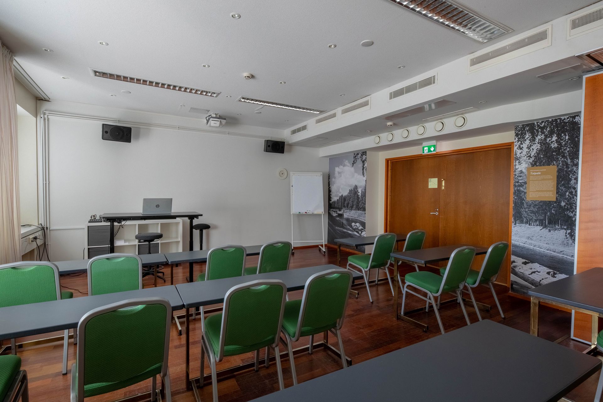 A meeting room with rows of green chairs facing a white board and a laptop on a desk, set against wood paneled walls.