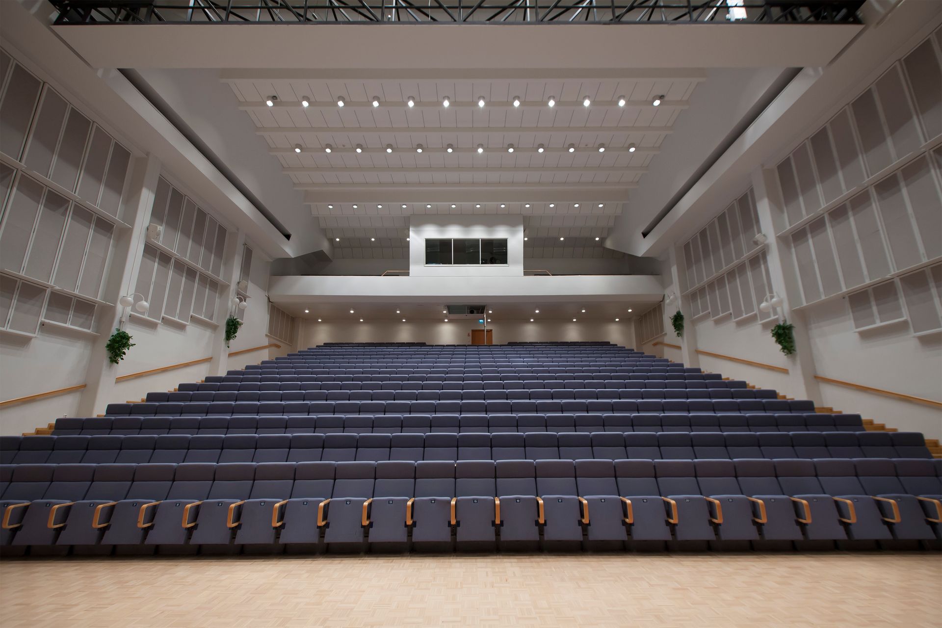 Empty lecture hall or auditorium with rows of blue tiered seats facing a central stage area under recessed lighting.