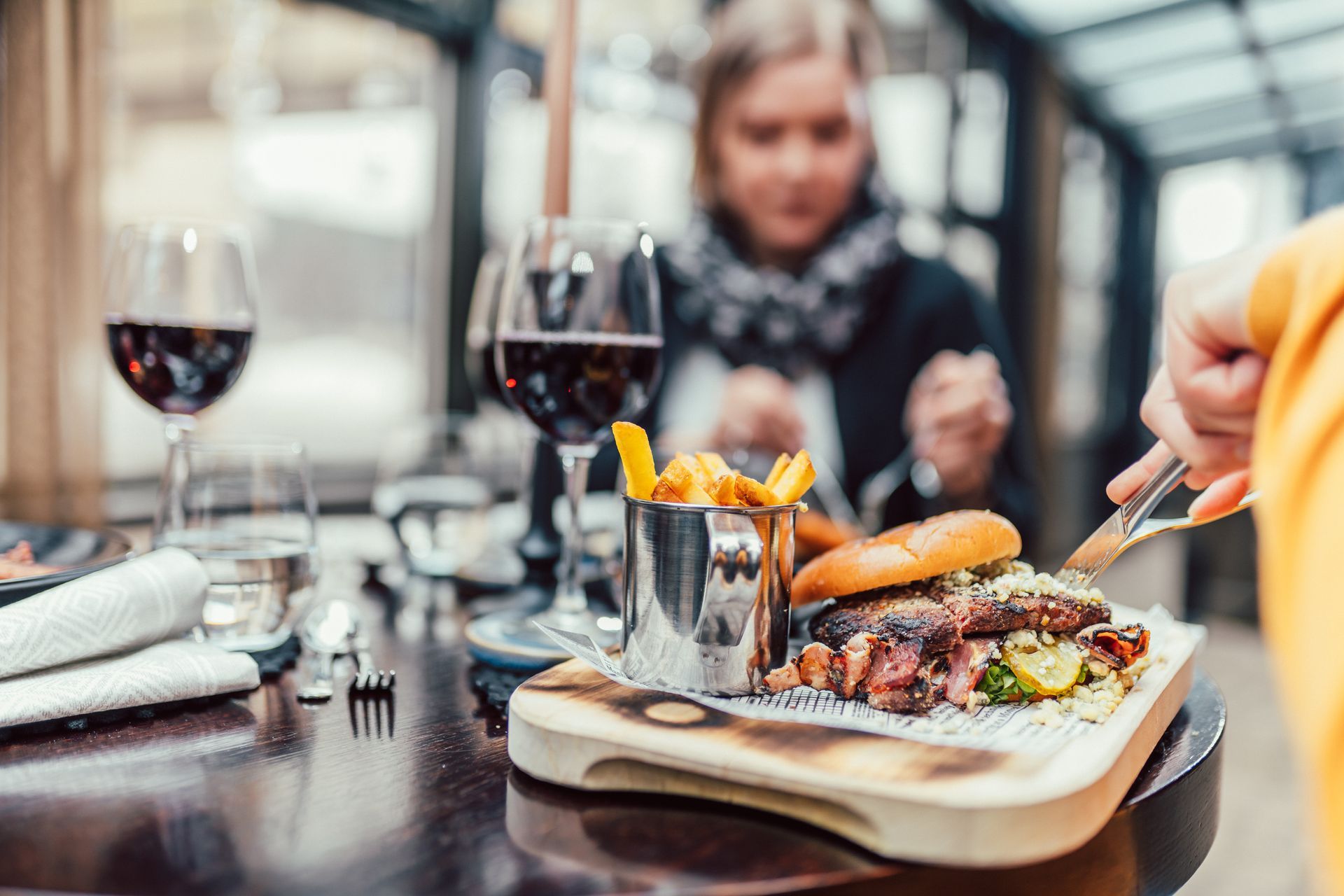 Henkilö leikkaa hampurilaista, joka tarjoillaan puulaudalla metallisen ranskanperunoiden kupin vieressä, taustalla viinilasit ja henkilö.